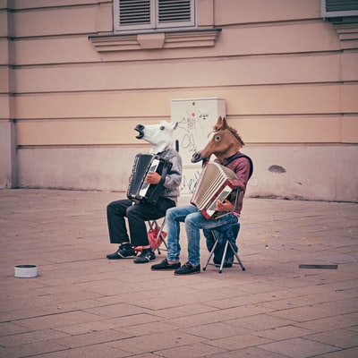 两个戴着马头的人坐在折叠椅上，在棕色的水泥建筑旁拉手风琴